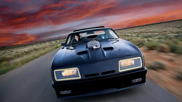 Max Rockatansky driving the Ford Falcon Interceptor on a highway in a post-apocalyptic setting.